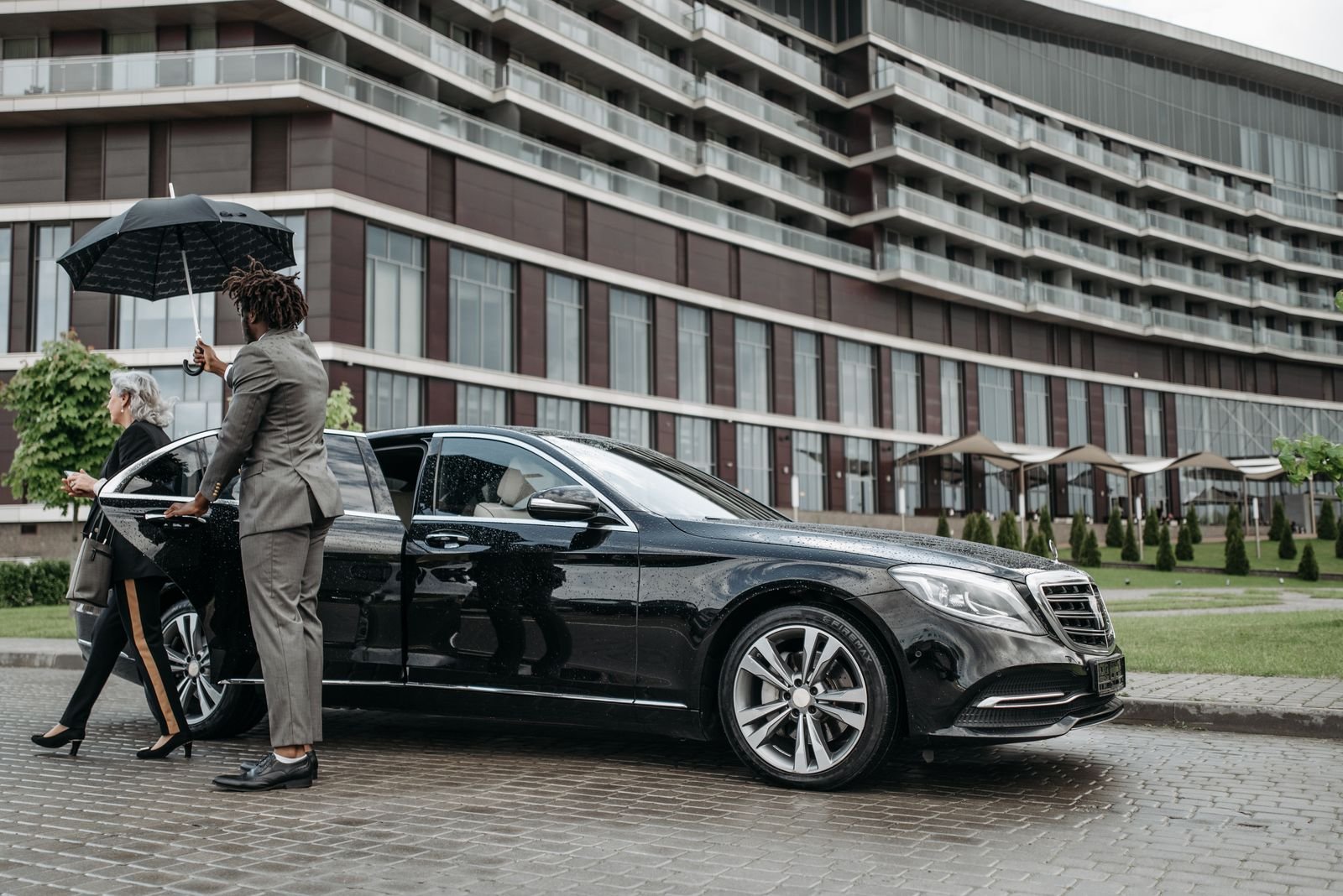 Lux4Rides chauffeur assisting a VIP passenger into a black Mercedes S-Class with an umbrella